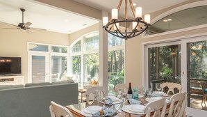 A dining area with a glass table, white chairs, and a chandelier. There is a large window with a view of greenery outside.