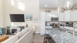 A modern kitchen with white cabinets, a granite countertop, and stainless steel appliances. There is a flat-screen TV mounted on the wall, a comfortable seating area with a sofa and a coffee table, and a dining area with a bench. - Luxury Beach Home | Heated Pool | Pets OK (Hilton Head Island)