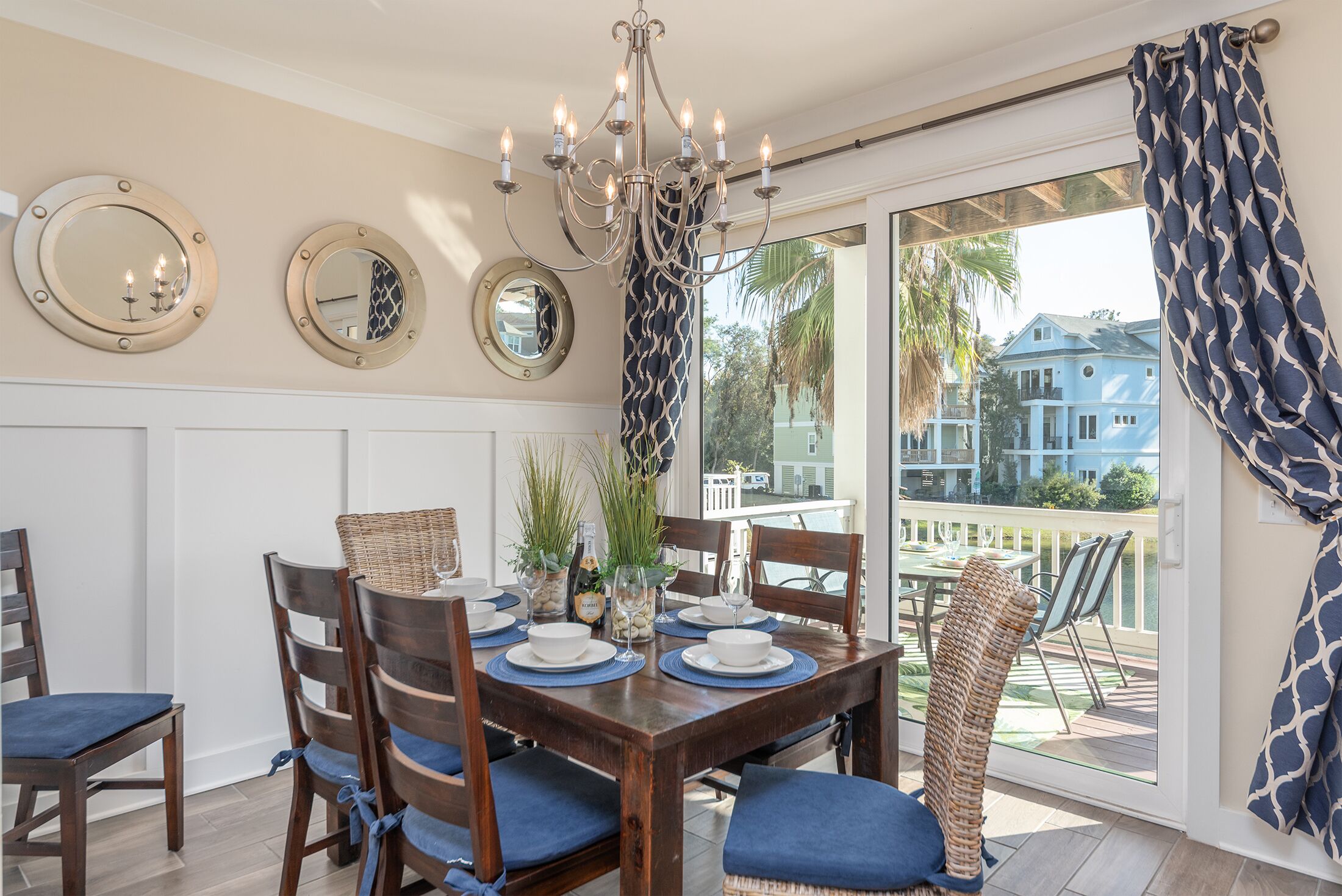A dining area with a wooden table set for six, surrounded by wicker chairs with blue cushions. There are three wall-mounted mirrors and a chandelier above the table.