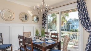 A dining area with a wooden table set for six, surrounded by wicker chairs with blue cushions. There are three wall-mounted mirrors and a chandelier above the table. - Luxury Beach Home | Heated Pool | Pets OK (Hilton Head Island)