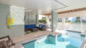 A pool area with a blue sofa, a surfboard sign, and a view of the neighborhood. - Luxury Beach Home | Heated Pool | Pets OK (Hilton Head Island)