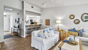 Uma sala de estar ampla com um sofá branco, uma mesa de centro de madeira e uma área de cozinha ao fundo.