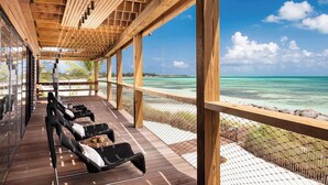 A covered outdoor seating area with wooden flooring and black wicker chairs, overlooking a beach with clear turquoise water and a distant shoreline. - Silver Sands Villa Turks and Caicos – 8-Bedroom Luxury Beachfront Villa with Gym & Padel Court (Thompson Cove)
