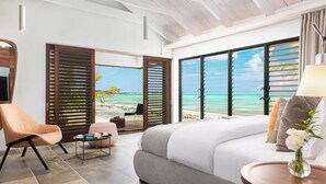 A hotel room with a large bed, a chair, a small table, and a view of the beach and ocean through open shutters. - Silver Sands Villa Turks and Caicos – 8-Bedroom Luxury Beachfront Villa with Gym & Padel Court (Thompson Cove)