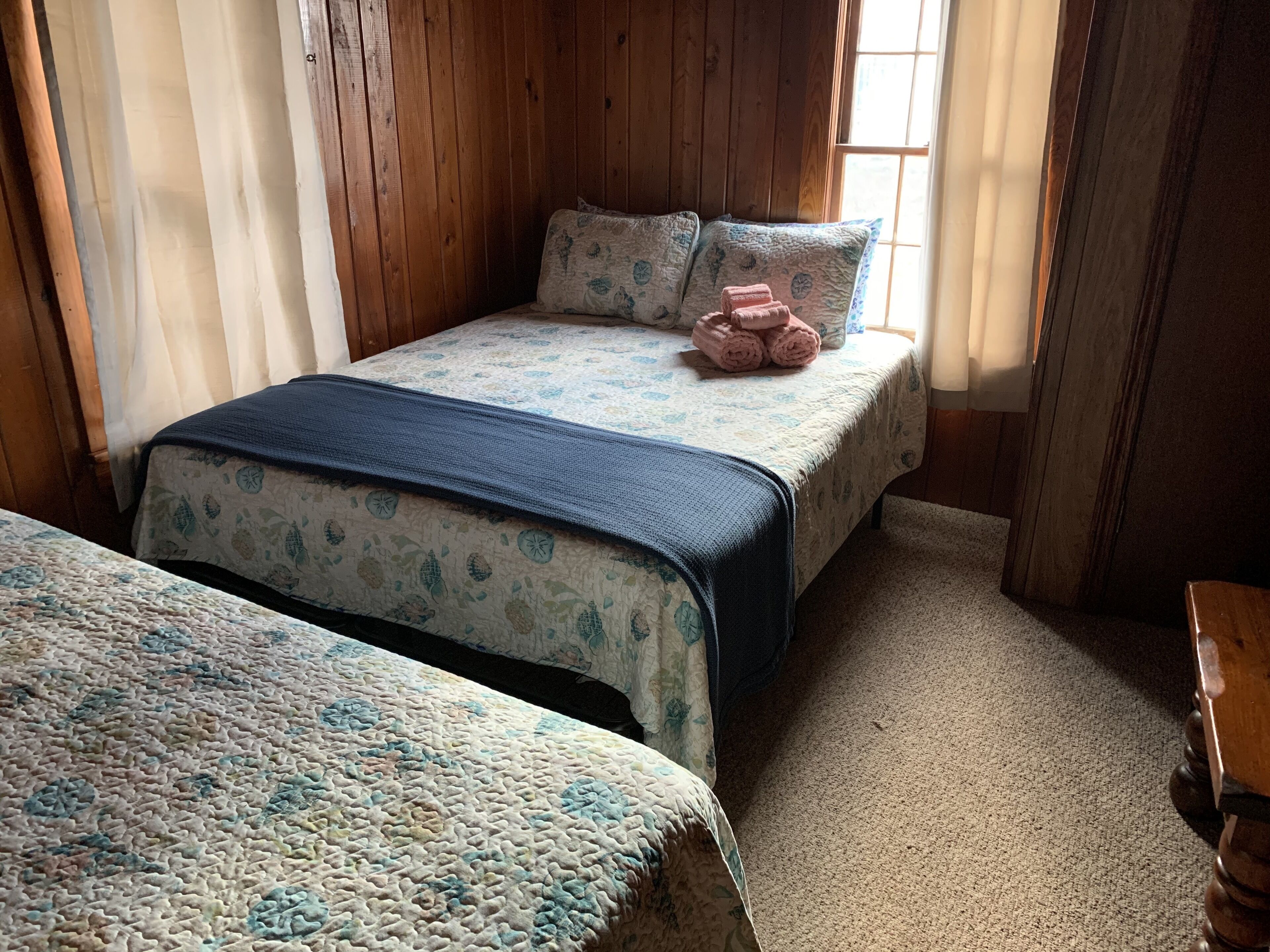 A room with two beds, wooden walls, a window with blinds, and a carpeted floor.