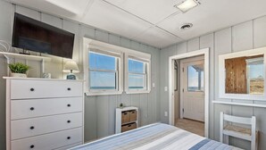 A bedroom with a bed, dresser, TV, windows, and a door leading to another room.