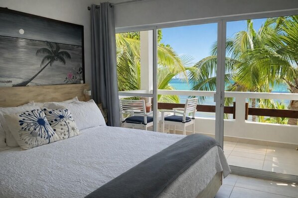A hotel room with a large bed, a painting of a palm tree, a balcony with a view of palm trees and the sea.