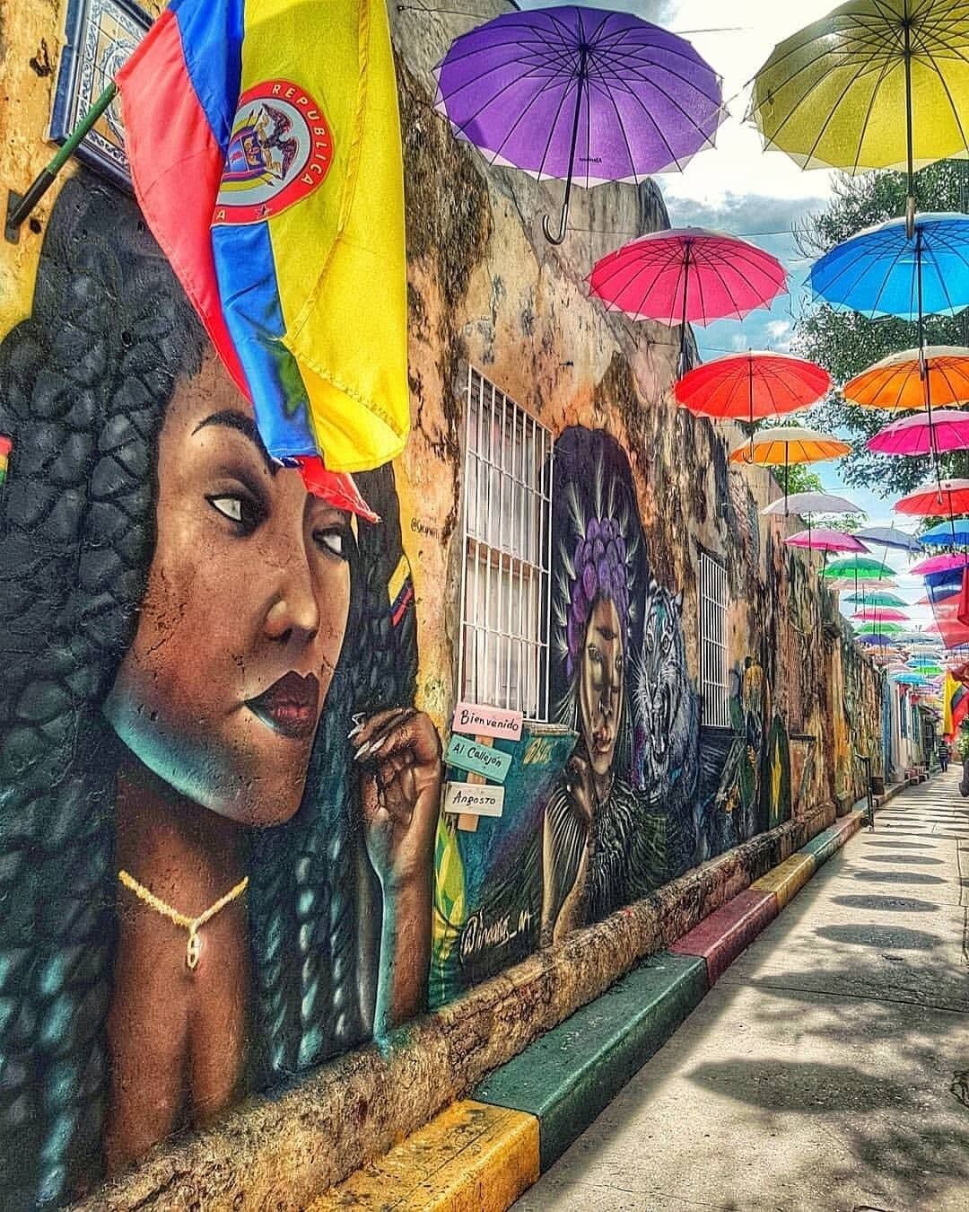 Un murale raffigura una persona che spia da un davanzale, ombrelloni colorati sopra e una strada con marciapiede.