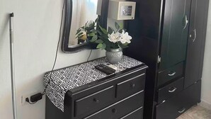 Una habitación con televisión montada en la pared, cómoda con cajones, espejo y armario.