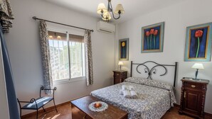 Un dormitorio con cama, una silla, una mesita y un ventanal con cortinas.