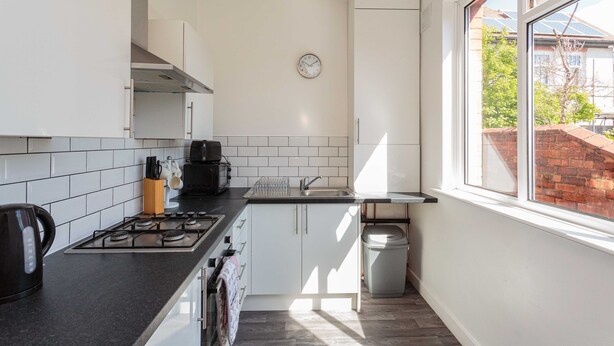 A modern kitchen with white cabinets, a gas stove, a coffee maker, and a window with a view of a brick building. - Cherry Property - Mulberry Suite (Blackpool)