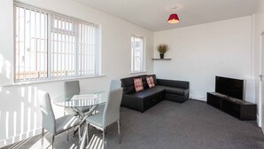 A modern living room with a glass table, two chairs, a black sectional sofa, a flat-screen TV, and a red pendant light. - Cherry Property - Mulberry Suite (Blackpool)