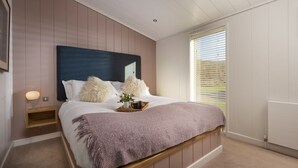 A bedroom with a large bed, a nightstand, a window with blinds, and a radiator. - Charterhouse, Strawberryfield Park -  a lodge that sleeps 2 guests  in 1 bedroom (Cheddar)