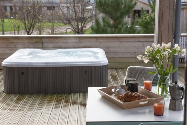 Un bain à remous sur une terrasse en bois, avec une table à proximité, sur laquelle se trouve un plateau avec des croissants, une théière et des verres de jus de fruits.