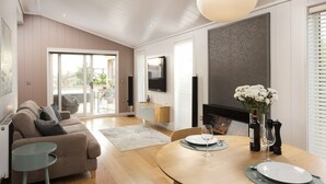 A modern living room with a sofa, dining table, and a TV mounted on the wall. - Charterhouse, Strawberryfield Park -  a lodge that sleeps 2 guests  in 1 bedroom (Cheddar)