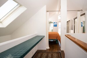 Un couloir étroit avec une lucarne, un lit recouvert d’une couverture orange et un banc en bois.
