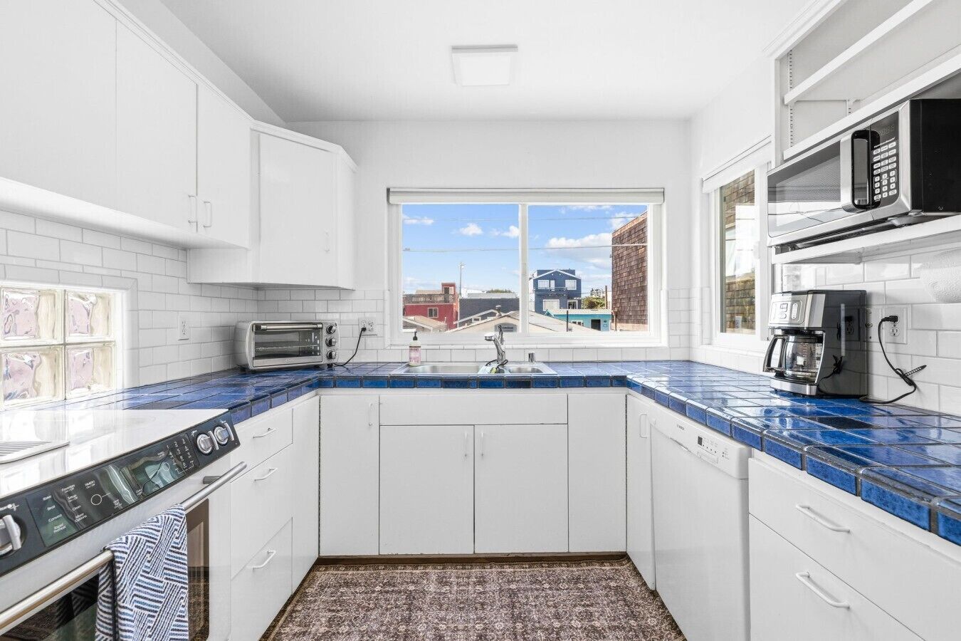 Eine moderne Küche mit weißen Schränken, einer blauen Fliesenspiegel, einem eingebauten Backofen, einer Kaffeemaschine und einem Fenster mit Blick auf Gebäude.