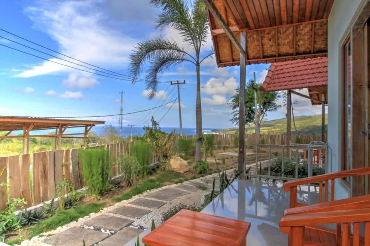 A wooden deck with a glass balustrade overlooking a garden with palm trees and a view of the ocean.