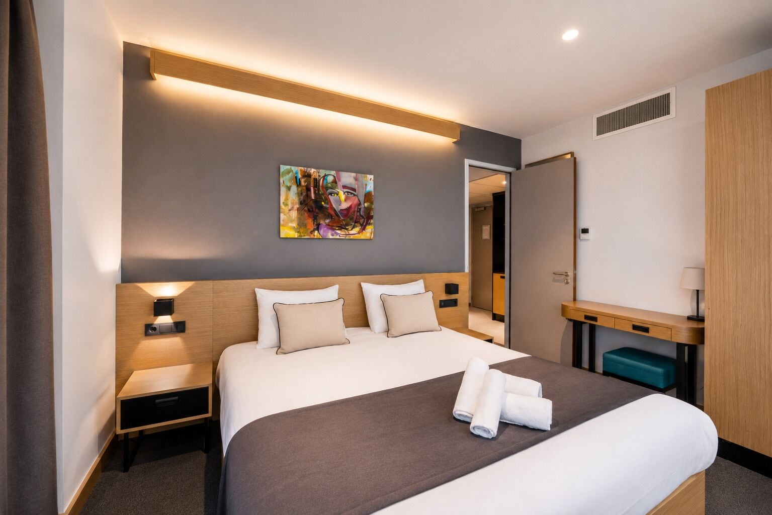 A hotel room with a large bed, bedside tables, a desk, a mirror, and a painting on the wall.