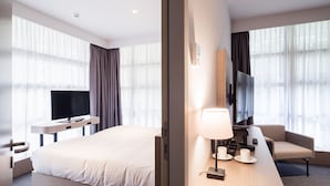 A modern hotel room with a large bed, a desk with a lamp, a television, and a seating area with a chair and a table.