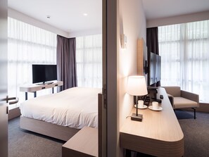 Une chambre d’hôtel moderne dotée d’un grand lit, d’un bureau avec une lampe, d’une télévision et d’un coin salon avec un fauteuil et une table.