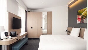 A modern hotel room with a large bed, a desk with a flat-screen TV, a bench, and a wardrobe.