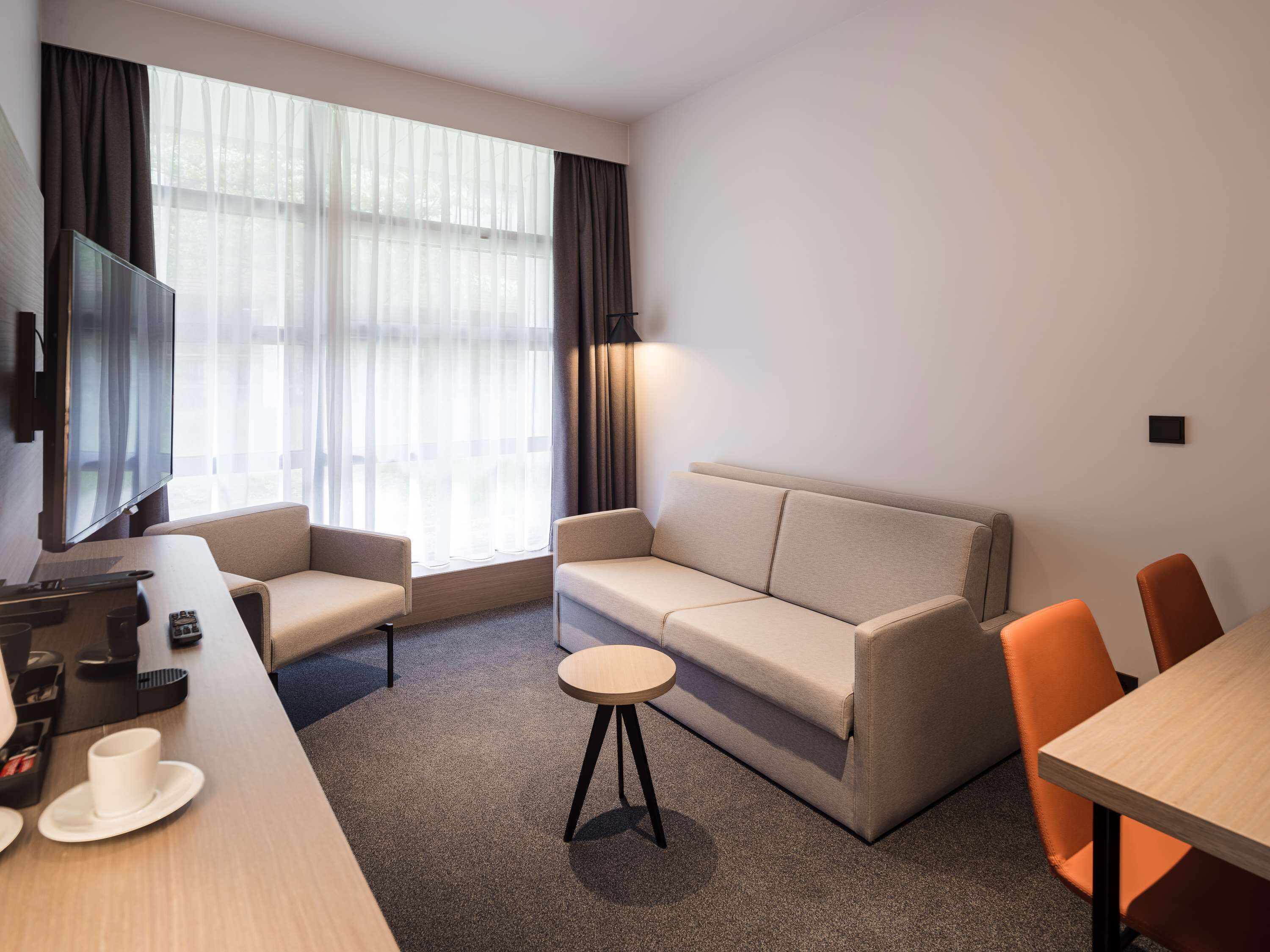 A modern hotel room with a sofa, two armchairs, a small round table, a flat-screen TV mounted on the wall, and a large window with sheer curtains.