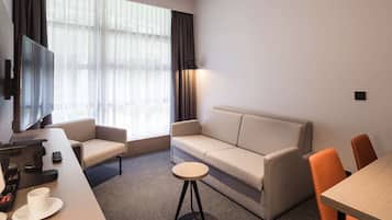 A modern hotel room with a sofa, two armchairs, a small round table, a flat-screen TV mounted on the wall, and a large window with sheer curtains.
