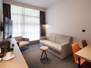 A modern hotel room with a sofa, two armchairs, a small round table, a flat-screen TV mounted on the wall, and a large window with sheer curtains. - Executive Residency by Best Western Amsterdam Airport (Hoofddorp)