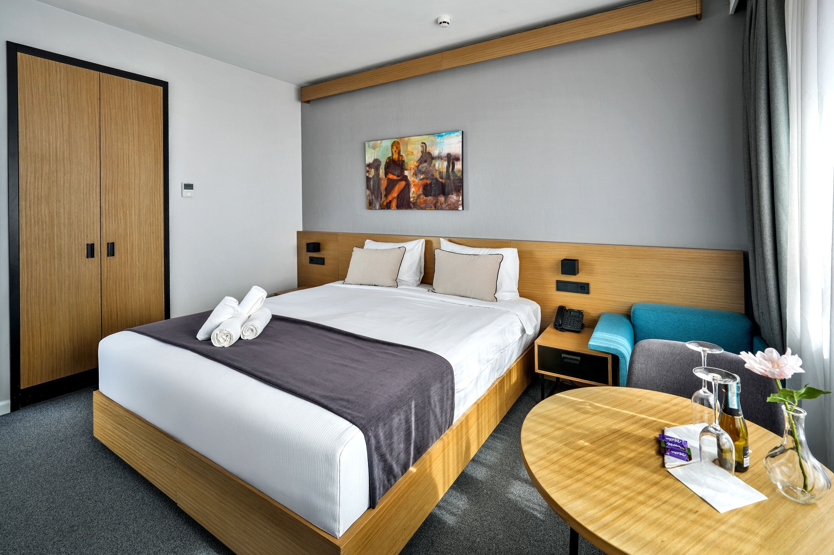 A hotel room with a bed, a bedside table with a phone and a bottle, a painting on the wall, and a window with curtains.
