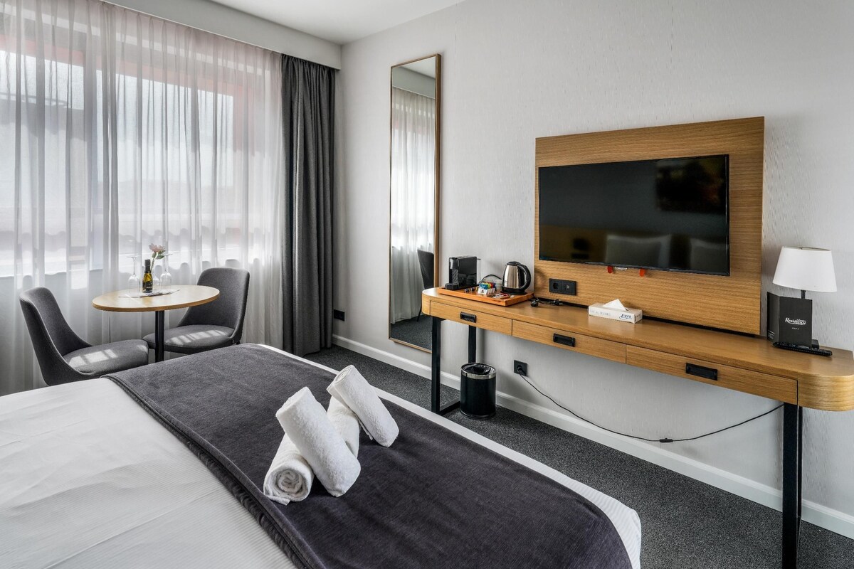 Una habitación de hotel con cama, escritorio, silla, televisor y ventana con cortinas.
