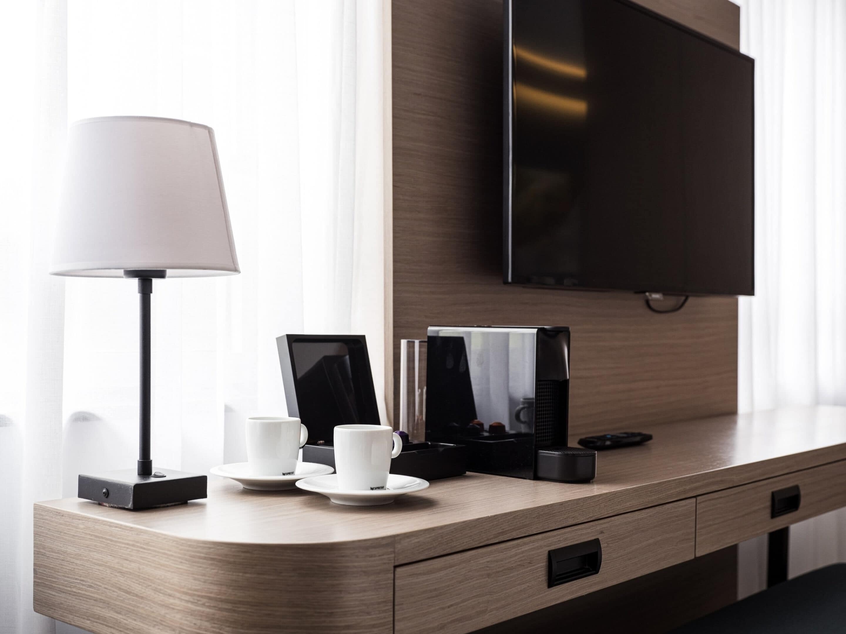 Habitación de hotel con un escritorio de madera, televisor de pantalla plana, lámpara y cafetera.