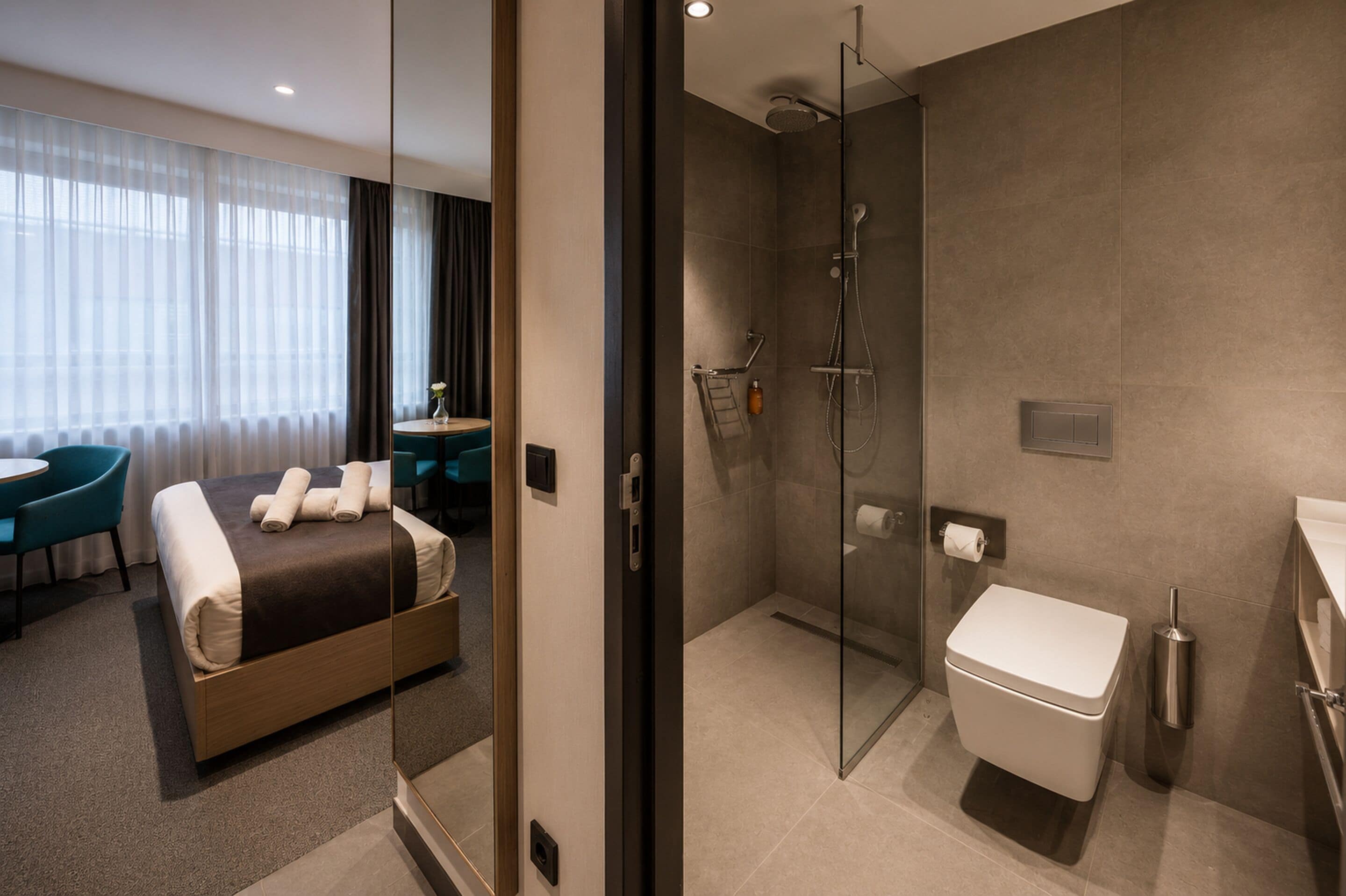 Ein modernes Hotelzimmer mit einer gläsernen Duschkabine, einem Bett mit ordentlich gefalteten Handtüchern, einem kleinen Tisch mit einer Vase und einer Sitzecke mit einem Sessel.