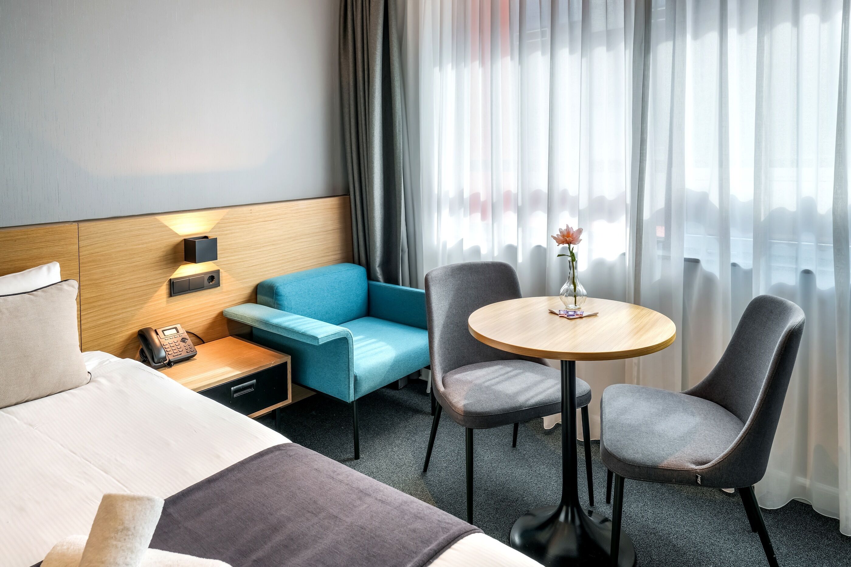 Habitación de hotel con cama, escritorio con teléfono, sofá azul, dos sillas, una mesita con un florero y una ventana con cortinas.