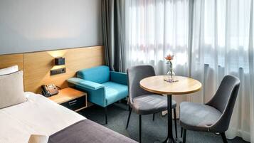Habitación de hotel con cama, escritorio con teléfono, sofá azul, dos sillas, una mesita con un florero y una ventana con cortinas.