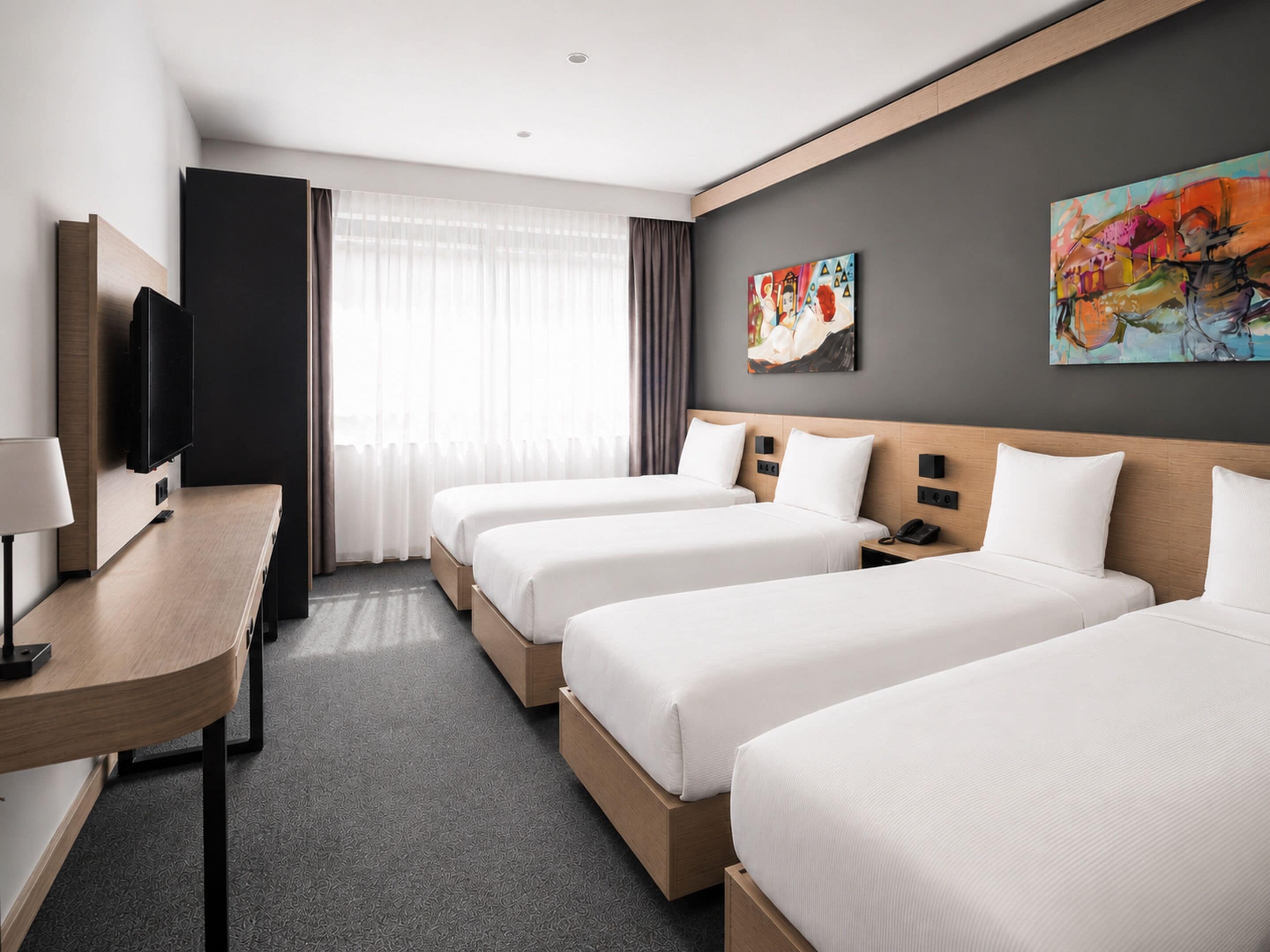 A hotel room with three beds, a desk, a flat-screen TV, and artwork on the walls.