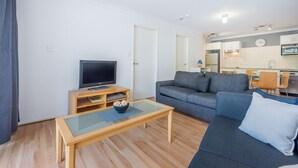 Un salon moderne avec un canapé, une table basse, un téléviseur posé sur un meuble et une cuisine en arrière-plan.