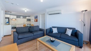Un salon moderne avec un canapé et une table basse, une cuisine avec des meubles blancs et un four à micro-ondes, et un climatiseur fixé au mur.