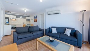 A modern living room with a sofa and a coffee table, a kitchen with white cabinets and a microwave, and an air conditioning unit on the wall.