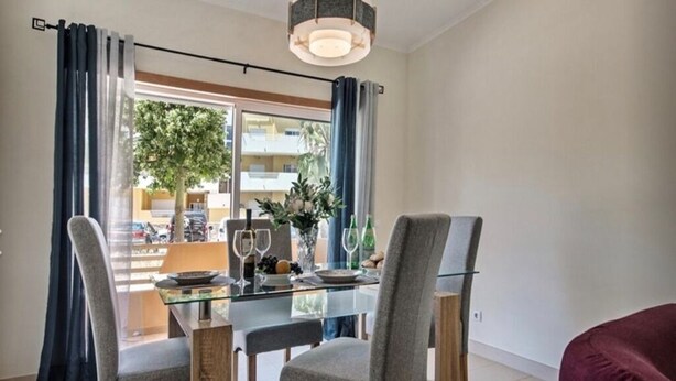 Comedor con mesa de vidrio, sillas tapizadas en gris y vista a un edificio y un árbol.