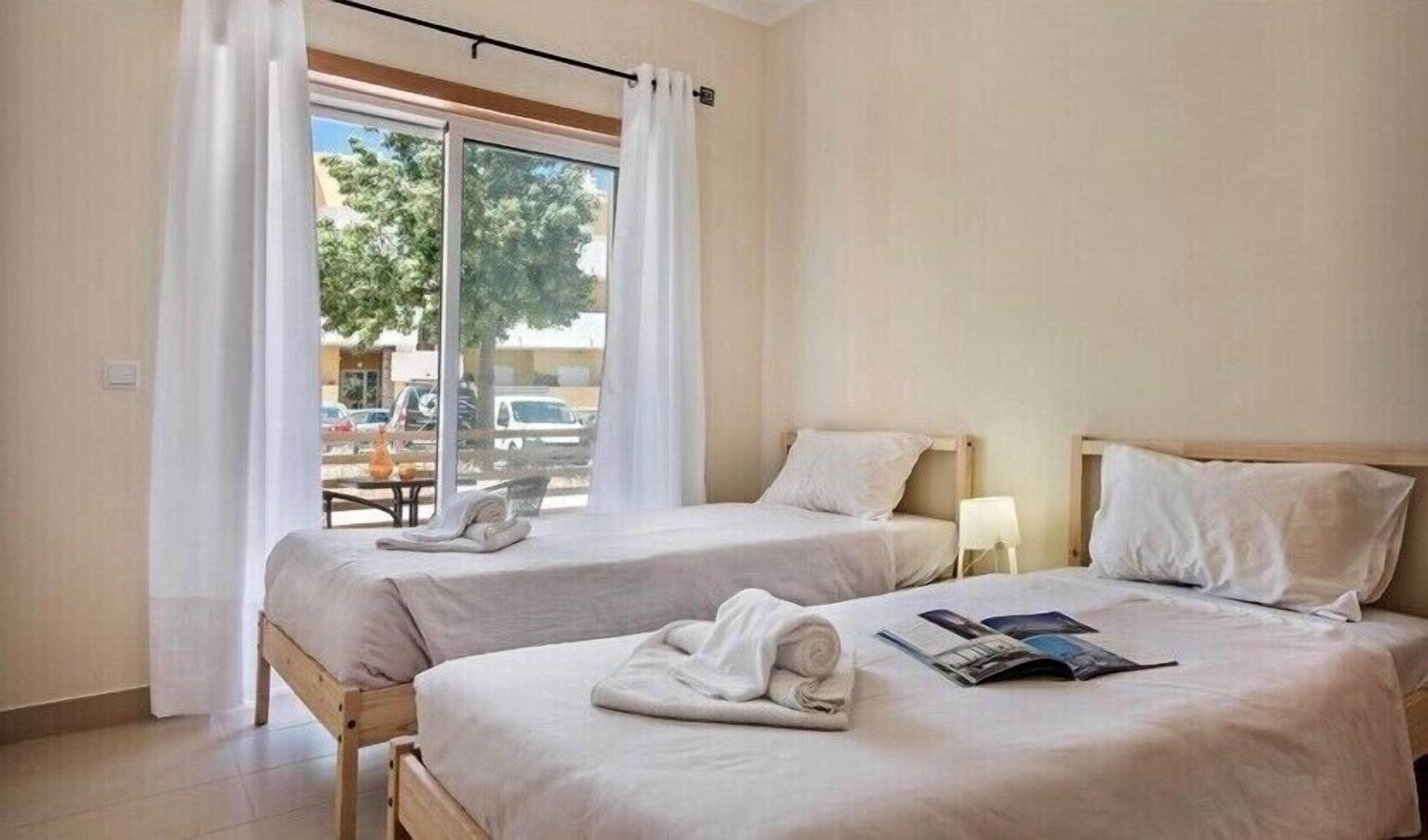 A hotel room with two beds, a window with curtains, a table with chairs, and a view of the outside.
