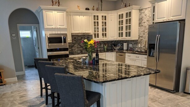 A modern kitchen with white cabinetry, a granite island with bar stools, a stainless steel refrigerator, and a brick backsplash. - Beautiful home on an 18 holes golf course (Sarasota)