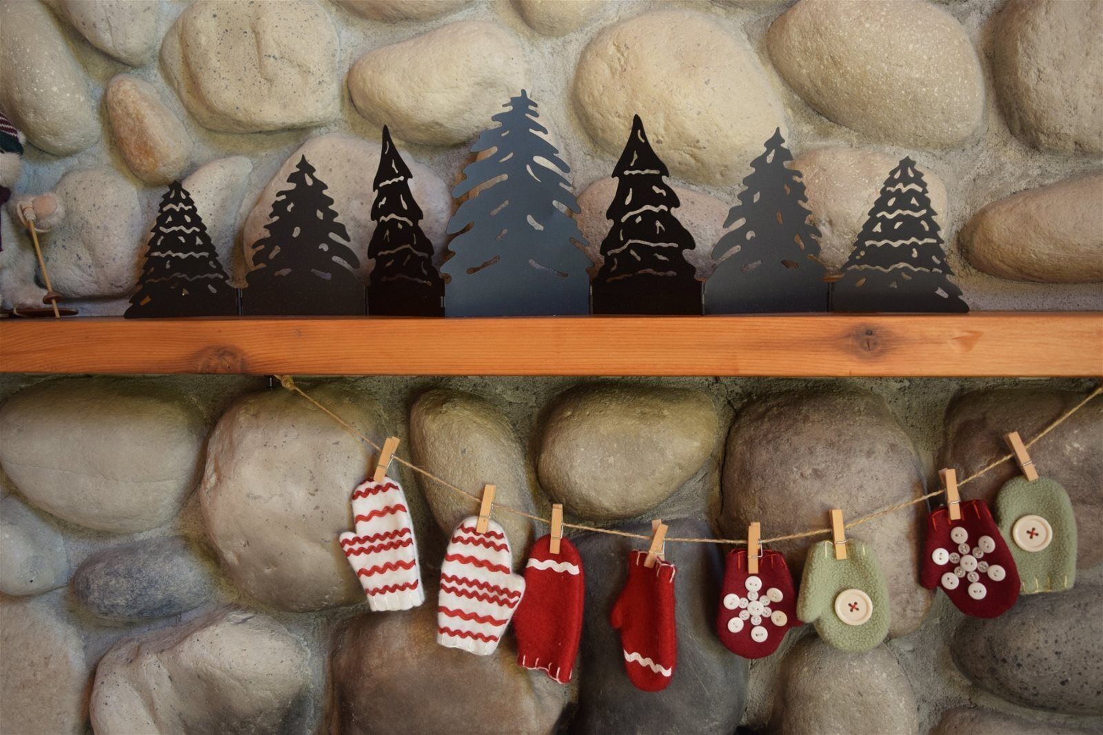 A wooden shelf with paper Christmas trees and mittens hanging on a string against a stone wall.