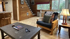 A cozy cabin interior with a wooden staircase, a dining area, a sofa with cushions, and a table with books and remotes.