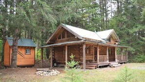 A rustic cabin with a porch, a small shed, and a fire pit in a forested area. - Cascade Log Cabin (Packwood)