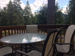 Een ronde glazen tafel met metalen stoelen op een houten terras, omringd door een houten balustrade en bomen.