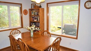 Une salle à manger avec une table et des chaises en bois, une bibliothèque et de grandes fenêtres donnant sur des arbres et sur un véhicule à l’extérieur.
