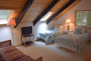 A cozy attic bedroom with a sloped ceiling, a television, two beds, a chair, and a lamp.