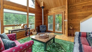 A cozy cabin interior with a large wooden ceiling, a fireplace, and a view of the forest through large windows. - Trails End (Packwood)