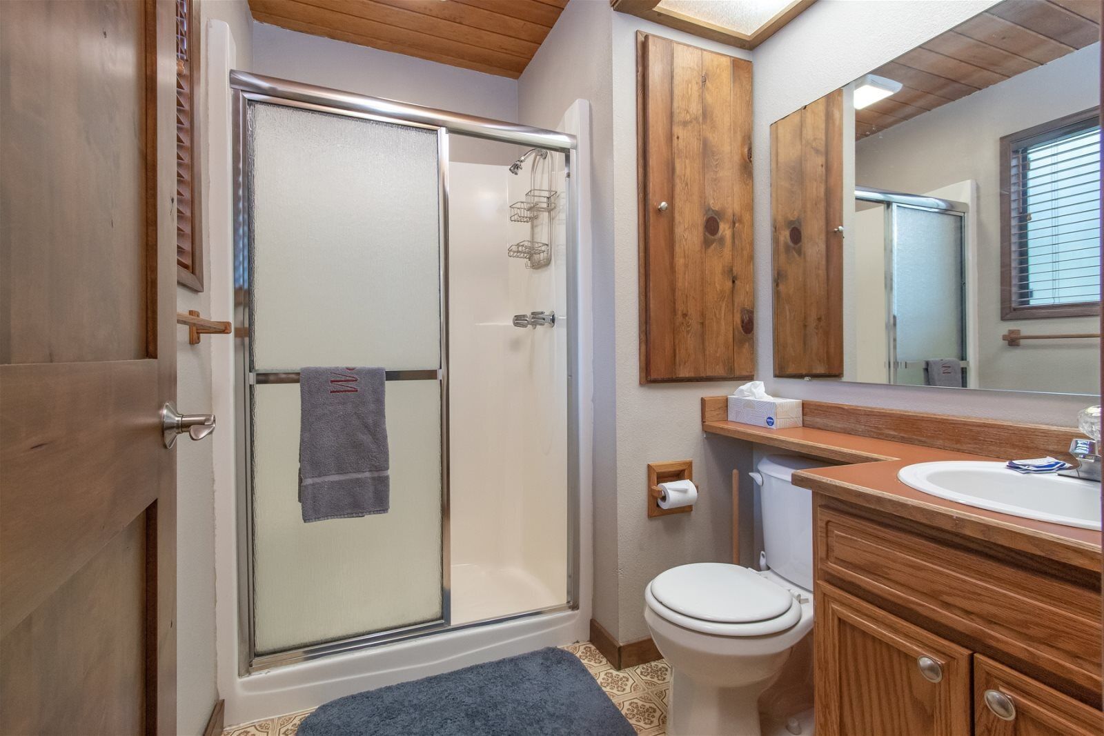 A bathroom with a glass shower enclosure, a toilet, a sink, and wooden cabinets.